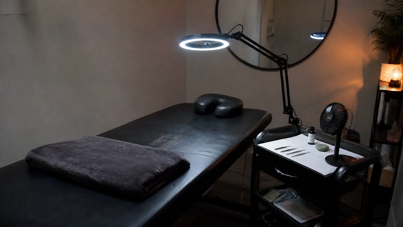 Lash extension room with a treatment bed, ring lamp on an arm and a rolling cart of tweezers and adhesive