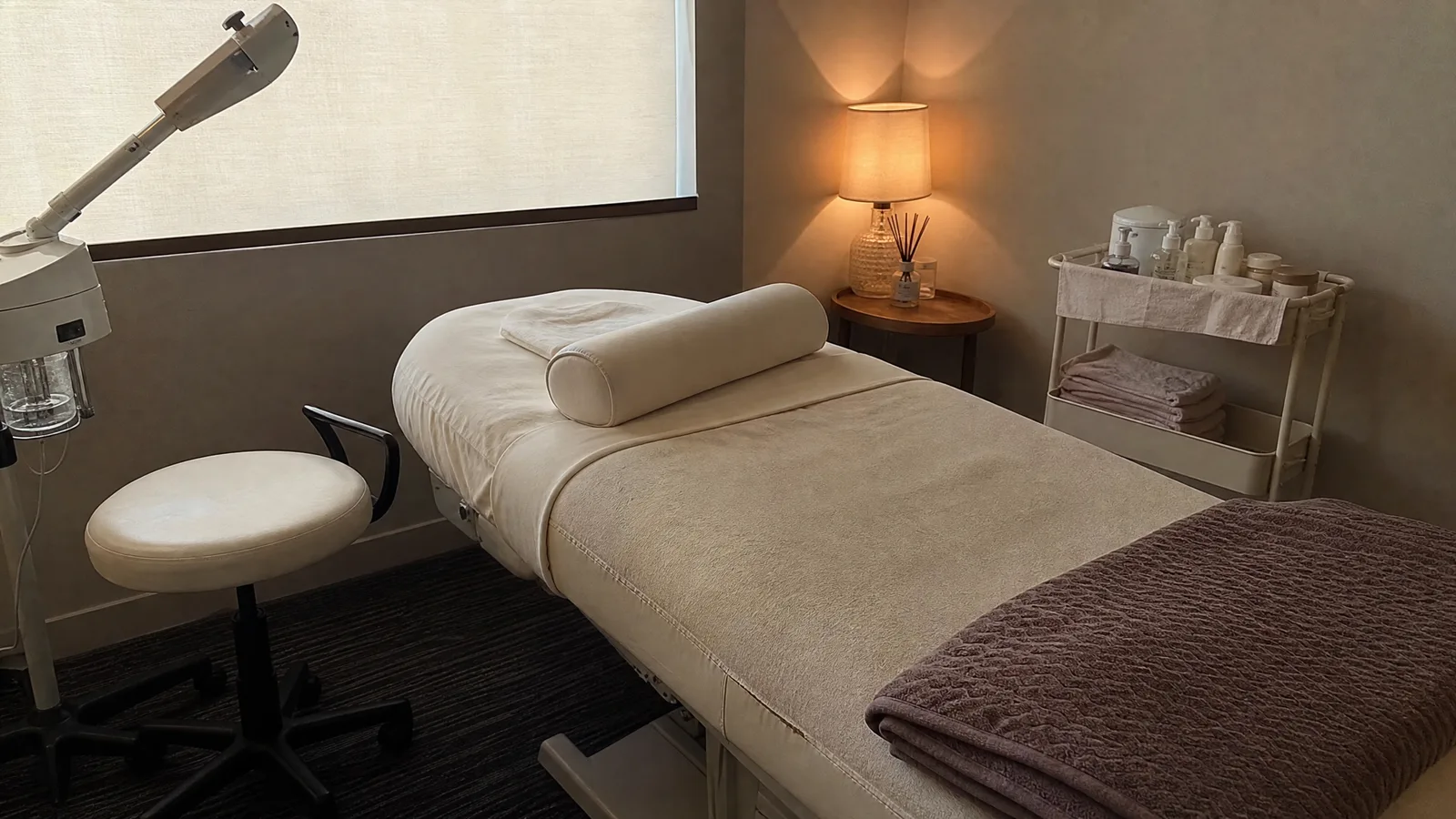 Spa treatment room with a draped table, rolling stool, steamer arm and a trolley of covered products